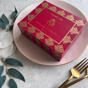 Puede incluir: Caja de regalo de boda roja y dorada con un s&iacute;mbolo Ganesha dorado y el texto "Gracias Shivayam & Babitha 10.02.24"