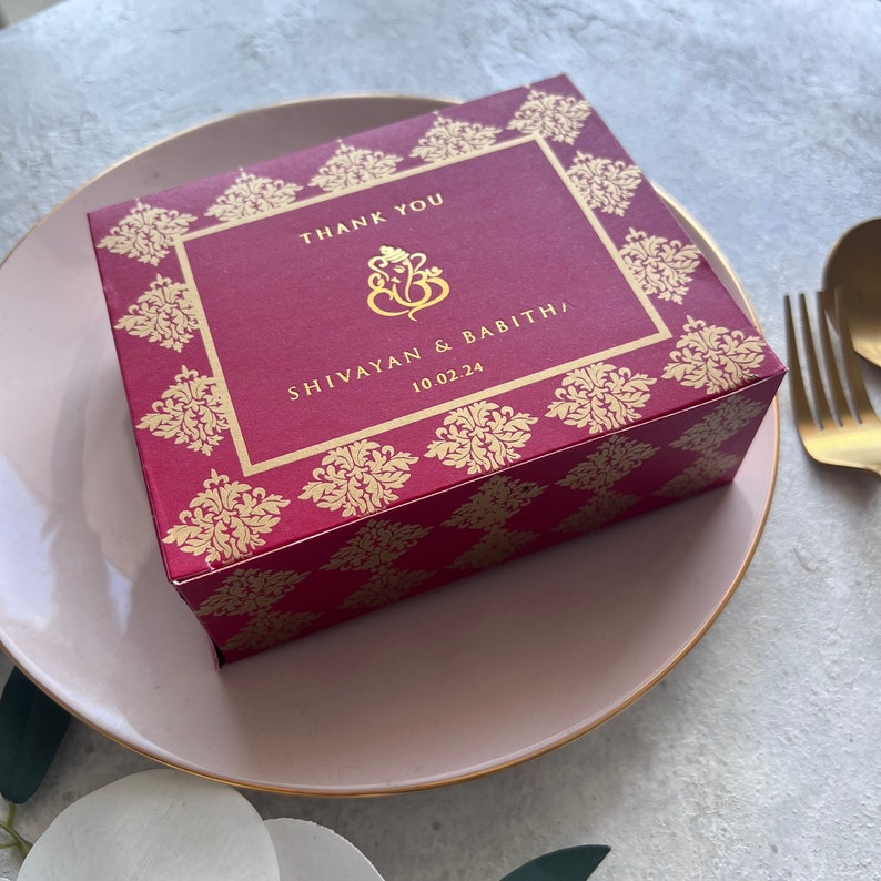 Puede incluir: Una caja de agradecimiento roja y dorada con un patr&oacute;n decorativo. La caja tiene el texto "Thank You" y "Shivayan & Babitha" con la fecha "10.02.24".
