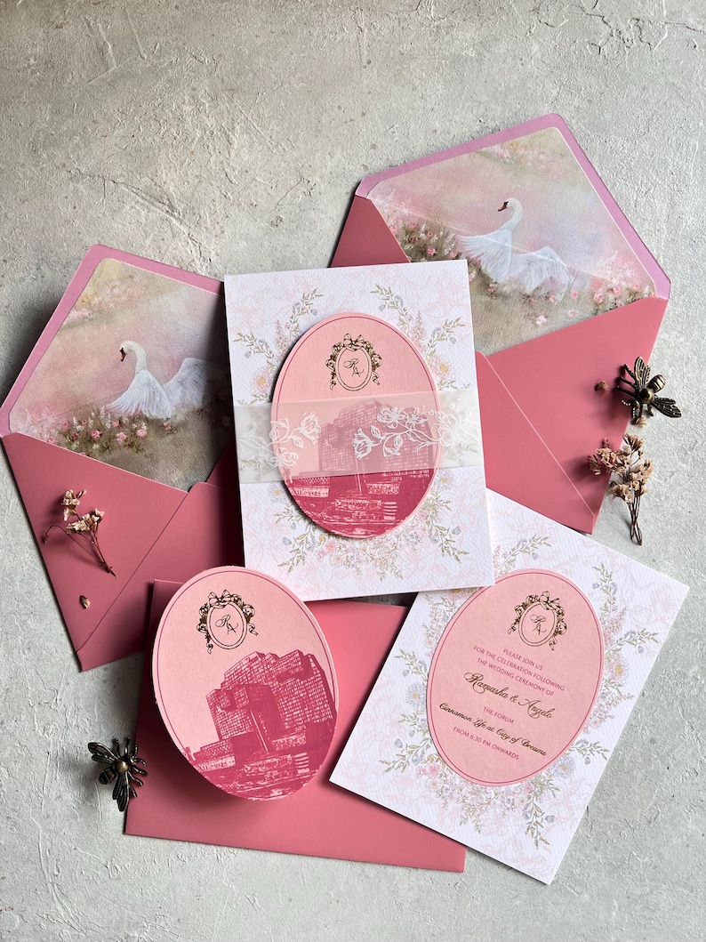 May include: Elegant wedding invitation suite in shades of pink and white. The set includes invitations, RSVP cards, and envelopes, adorned with floral patterns, swan illustrations, and a building graphic. The invitations feature the names "Ragaska & Angele".