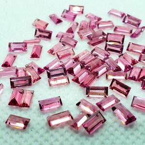May include: A collection of pink rectangular gemstones, likely tourmalines, scattered on a white surface. The stones are faceted and have a translucent appearance.