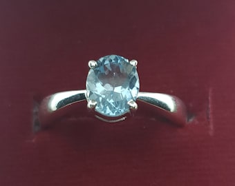 May include: A silver ring featuring a light blue oval gemstone. The gemstone is the focal point, set in a classic four-prong setting. The band is smooth and polished, with a simple, elegant design. The ring is displayed against a dark red background.