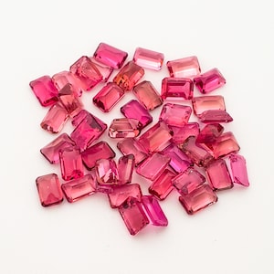 May include: A collection of pink, faceted, rectangular gemstones. The gemstones are scattered on a white surface.