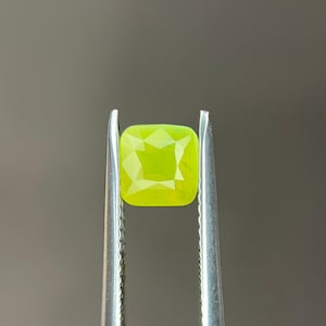 May include: A square-cut, vibrant, lime-green gemstone is held by silver tweezers against a gray background. The gemstone has a faceted surface, reflecting light. The overall theme is jewelry and gemstones.