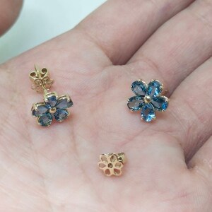 May include: A pair of gold flower-shaped earrings with blue gemstones. The earrings are on a hand.
