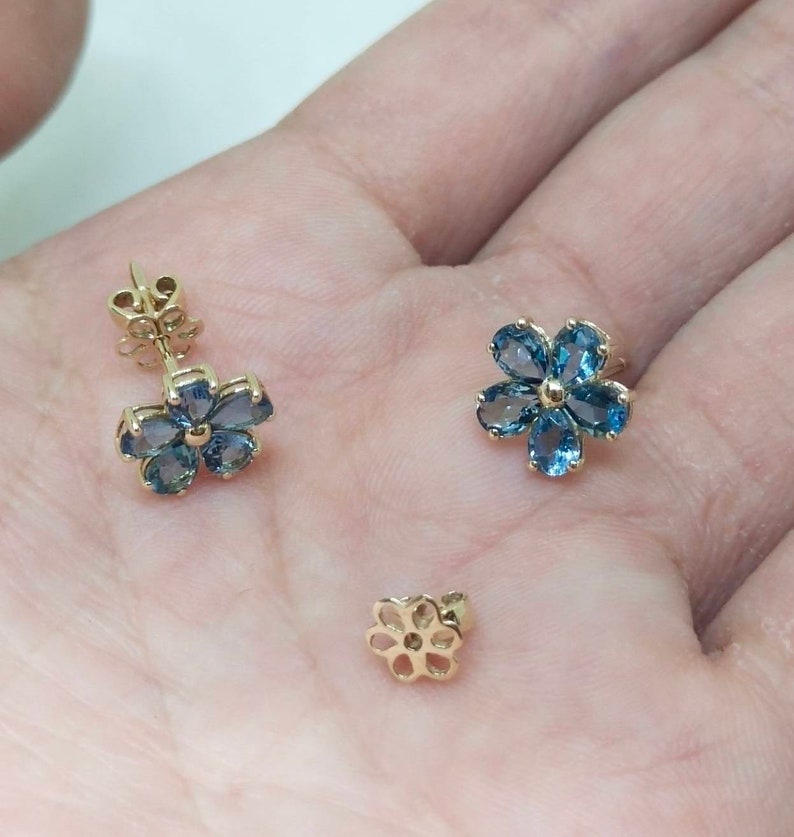 May include: A pair of gold flower-shaped earrings with blue gemstones. The earrings are on a hand.