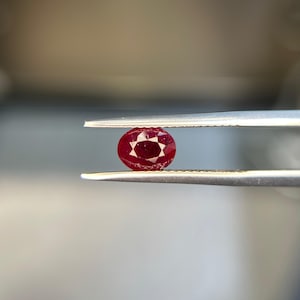 Puede incluir: Una gema de rubí ovalada y facetada sostenida por unas pinzas. El rubí es de color rojo intenso con un patrón en forma de estrella visible dentro de la piedra. Las pinzas son de color plateado y el fondo está borroso.