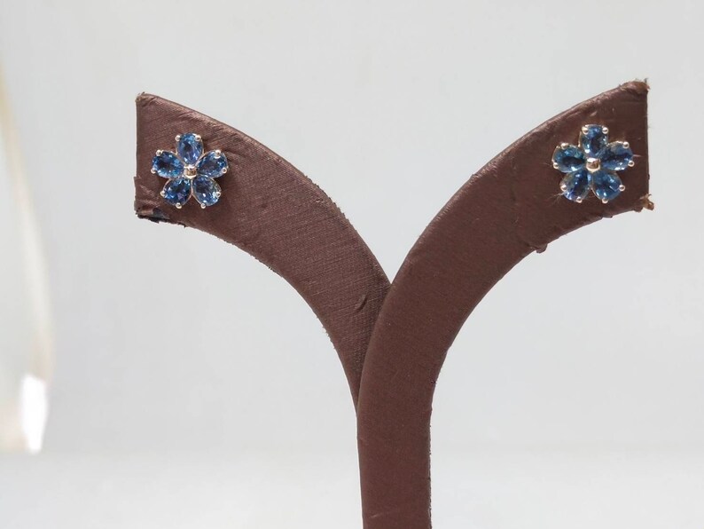 May include: A pair of blue sapphire flower stud earrings on a brown display stand.