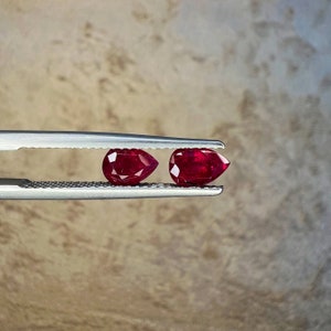 May include: Two pear-shaped ruby gemstones held in a pair of silver tweezers. The rubies are a deep red color and have a high shine.