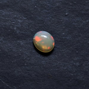 May include: A white opal gemstone with a light green and pink iridescent sheen. The oval-shaped stone is set against a dark gray background.
