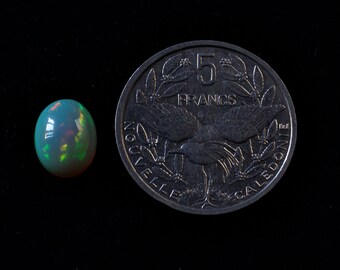 May include: An oval-shaped opal gemstone with iridescent colors of green, orange, and blue sits next to a silver coin. The coin is marked with the number 5 and the word FRANCS, with additional text around the edge.