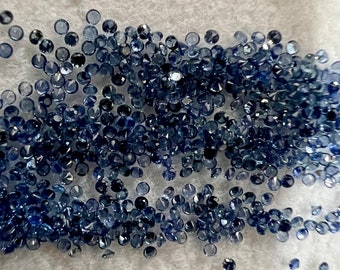 May include: A large quantity of small, dark blue sapphire gemstones scattered on a white surface.