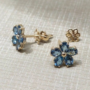 May include: A pair of gold flower-shaped earrings with blue gemstones. The earrings are on a white fabric background.