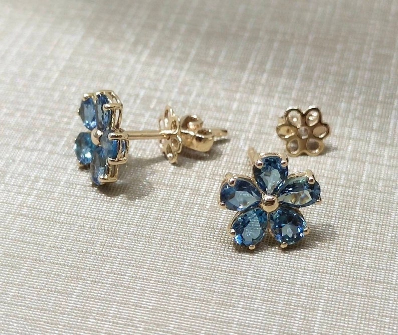 May include: A pair of gold flower-shaped earrings with blue gemstones. The earrings are on a white fabric background.
