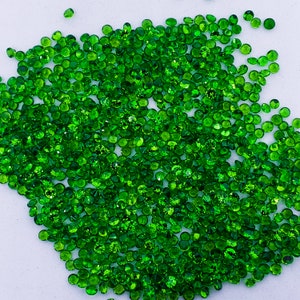 May include: A large group of small, round, green gemstones. The gemstones are scattered on a white surface.