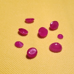 May include: A collection of eight pink gemstones, some oval shaped and some round, on a yellow surface.