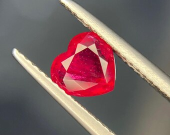 May include: A vibrant, heart-shaped red gemstone held securely by a pair of silver-toned metal tweezers. The faceted stone displays a brilliant crimson hue with visible internal light reflections and sharp, geometric cut edges throughout.