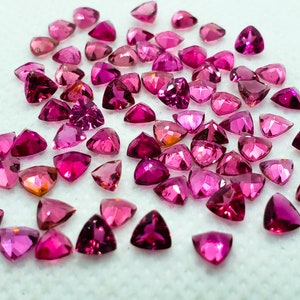 Könnte beinhalten: Eine Sammlung von dreieckigen rosa und roten Edelsteinen, auch bekannt als rosa Saphire, die auf einer weißen Oberfläche verstreut sind.