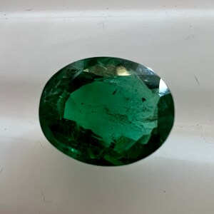 May include: A deep green oval-shaped gemstone with facets. The gemstone is on a white surface.