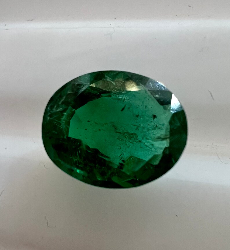 May include: A deep green oval-shaped gemstone with facets. The gemstone is on a white surface.