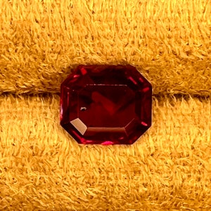 May include: A deep red gemstone with a faceted octagonal shape. The gemstone is set against a yellow, fuzzy background.