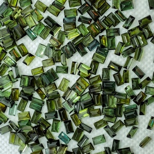 Op de afbeelding: Een verzameling losse groene edelstenen, gesneden in rechthoekige vormen, verspreid over een wit oppervlak.