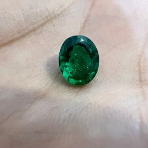 May include: A vibrant green emerald gemstone, cut into an oval shape, is displayed on a white surface.
