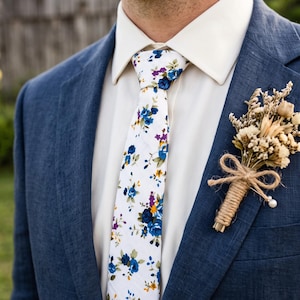 Könnte beinhalten: Ein marineblaues Sakko über einem cremefarbenen Hemd mit einer weißen Krawatte mit blauem, violettem und gelbem Blumenmuster. Ein Ansteckblümchen aus getrockneten Blumen, umwickelt mit Bindfaden, ist am Revers des Sakkos befestigt.