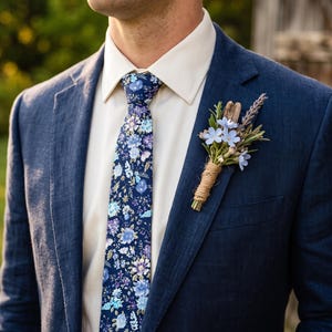 Op de afbeelding: Een marineblauw colbert gedragen met een crèmekleurig overhemd, een marineblauwe stropdas met een veelkleurig bloemenpatroon en een corsage bestaande uit lichtblauwe bloemen, groene takjes en een stuk drijfhout omwikkeld met touw.