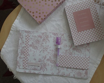 May include: A pink and white floral patterned notebook with a pink and white checkered pocket and a purple pen. The notebook has the text "perfectly imperfect" on the front.