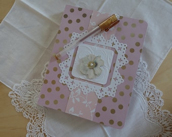 May include: A pink and gold polka dot journal with a white lace overlay and a delicate flower embellishment. A gold pen is lying on top of the journal.