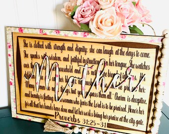 May include: A wooden sign with a floral background and a cutout of the word "Mother" in black. The sign features a quote from Proverbs 31:25-31, which reads: "She is clothed with strength and dignity; she can laugh at the days to come. She speaks with wisdom, and faithful instruction is on her tongue. She watches over the affairs of her household and is not afraid of the cold of winter. For her hands have done and let her works bring her praise at the city gate."