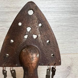 May include: A rustic, bronze-colored metal sculpture with a triangular top featuring several circular and irregular cutouts. The sculpture has a rounded base and several small, key-shaped metal charms hanging from the bottom of the triangle.