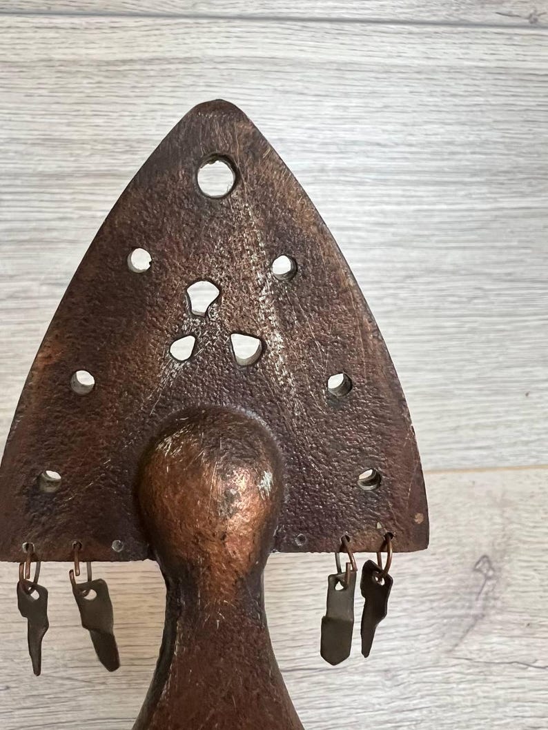 May include: A rustic, bronze-colored metal sculpture with a triangular top featuring several circular and irregular cutouts. The sculpture has a rounded base and several small, key-shaped metal charms hanging from the bottom of the triangle.