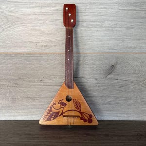 May include: A wooden balalaika, a traditional Russian stringed instrument, with a triangular body and a long neck. The instrument features a decorative design of a bird and berries in shades of red and brown. The balalaika is set against a wood-grain background.