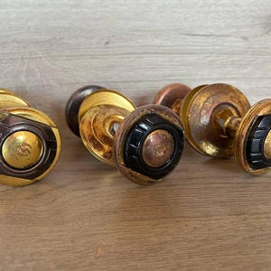 May include: A selection of antique brass door knobs with a gold and black finish. The knobs have a round shape with a decorative design and a small, circular emblem in the centre. The knobs are arranged on a wooden surface.