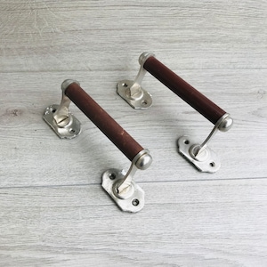 Może przedstawiać: Dwa srebrne uchwyty do drzwi szafki z brązowymi drewnianymi uchwytami. Uchwyty są zamontowane na jasnym drewnianym podłożu.