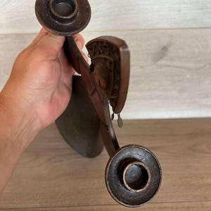 May include: A dark brown, antique candle holder with two circular candle cups. The holder has an ornate, carved design with small dangling metal accents. The piece is held in a hand, with a wooden surface in the background.