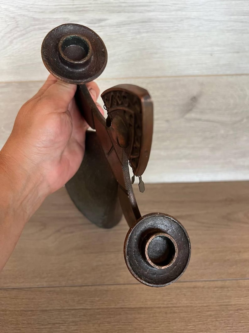 May include: A dark brown, antique candle holder with two circular candle cups. The holder has an ornate, carved design with small dangling metal accents. The piece is held in a hand, with a wooden surface in the background.