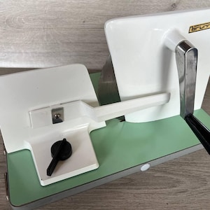 May include: Vintage white and green Brunner meat slicer. The slicer has a white handle, a black handle, and a black knob. The base is green with a silver trim. The word "Brunner" is printed in a gold rectangle.