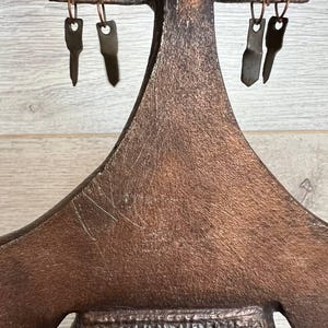 May include: A handcrafted, bronze-colored metal key holder with a unique, abstract design. The key holder features a textured surface and several key-shaped hooks for hanging keys. The design includes a central figure with outstretched arms and a textured lower section.