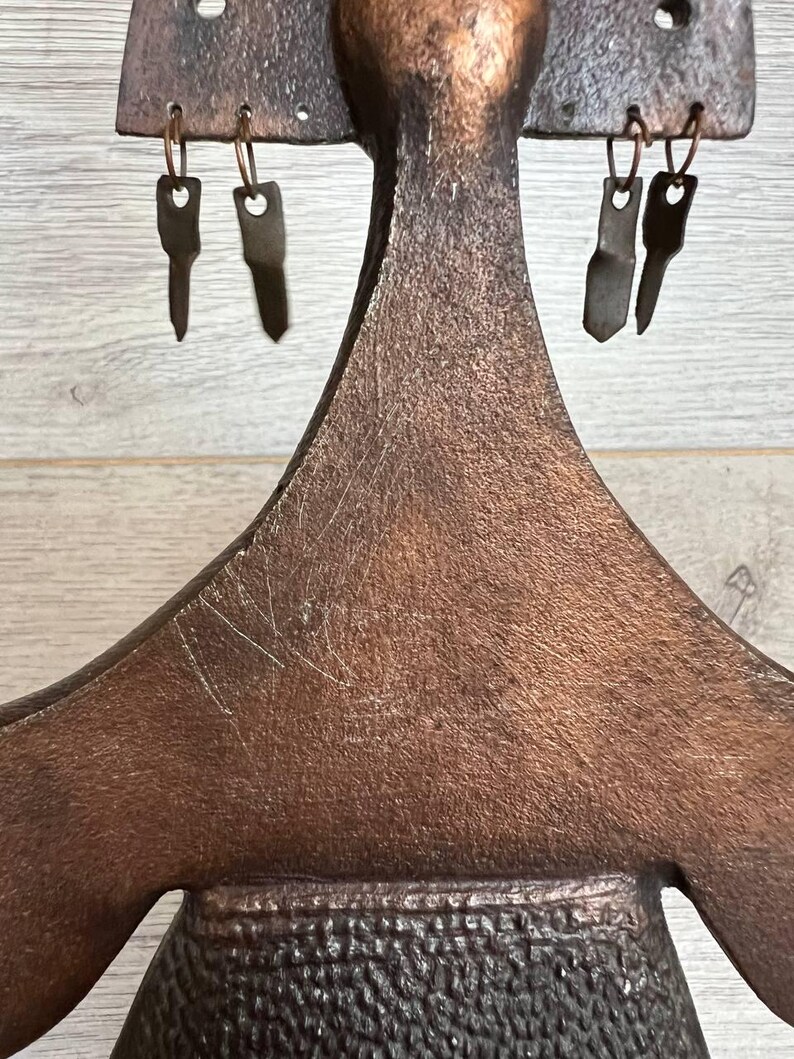 May include: A handcrafted, bronze-colored metal key holder with a unique, abstract design. The key holder features a textured surface and several key-shaped hooks for hanging keys. The design includes a central figure with outstretched arms and a textured lower section.