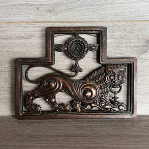 May include: A bronze wall sculpture featuring a stylized lion with intricate details. The lion is framed by a decorative border with a floral design.