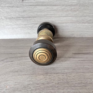 May include: A black and brass door knob with a circular design. The knob has a series of concentric circles in a brass finish.