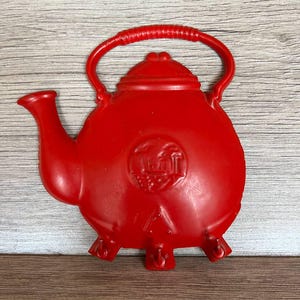 Pode incluir: Decoração de parede em forma de chaleira vermelha brilhante. A chaleira tem um corpo arredondado, um bico curvo e uma alça em forma de laço. O design inclui uma tampa e três pés pequenos. Um emblema circular está na frente.