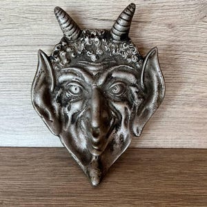 Könnte beinhalten: Ein silberfarbener Wandbehang mit Teufelsgesicht, Hörnern, spitzen Ohren und detailliertem Gesichtsausdruck. Die Skulptur hat eine strukturierte Oberfläche und ist auf einem Holzgrund befestigt. Das Kunstwerk ist ein dekoratives Stück.