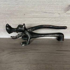 Può includere: Un apribottiglie in metallo nero con un design a forma di cane. L'apribottiglie ha un lungo manico e una testa dettagliata con mascelle aperte. L'oggetto è posto su una superficie di legno.