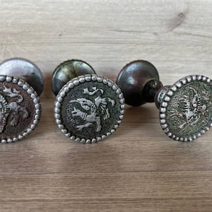 May include: Three round metal drawer knobs with a dark, weathered finish. Each knob features a raised, silver-toned relief of a winged griffin creature in the center, surrounded by a beaded border. The knobs have a cylindrical base and a textured surface.