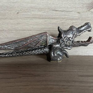 May include: A silver dragon nutcracker with an open mouth and detailed wings. The nutcracker is made of metal and has a rustic finish.