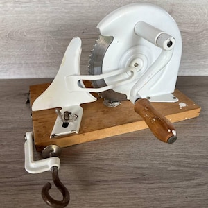 May include: A vintage white meat slicer with a circular saw blade, mounted on a wooden base. It features a wooden handle and a clamp for securing it to a surface. The slicer is designed for manual operation.