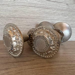 Könnte beinhalten: Zwei antike, silberfarbene Türknöpfe mit aufwendigen, geprägten Details. Jeder Knopf hat ein kreisförmiges Design mit einer zentralen Scheibe und floralen Mustern. Die Knöpfe sind durch eine Metallstange verbunden.